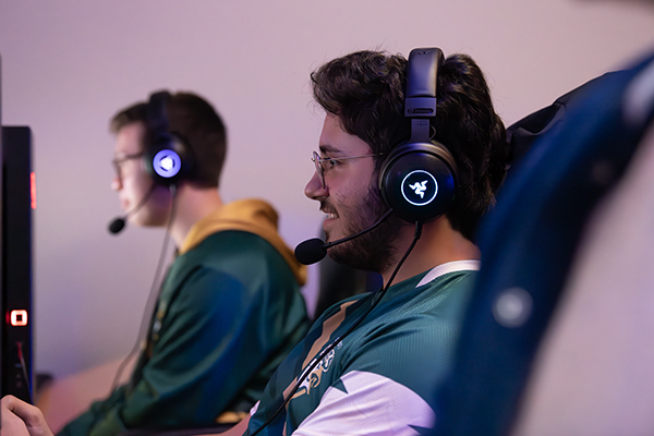 Two students wearing headsets and Husson Esports uniforms sit playing video games.
